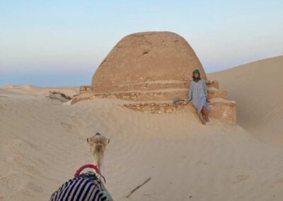 Kuppelförmiges Häuschen in der Wüste, fast im Sand versundken mit Frau und Kamel davor