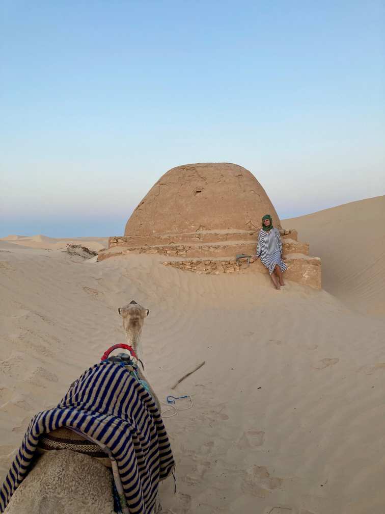 Kuppelförmiges Häuschen in der Wüste, fast im Sand versundken mit Frau und Kamel davor