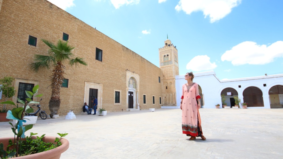 Eine Frau steht auf einem Hof in einer orientalischen Stadt, vor einem Gebäude, dahinter ein Minarett